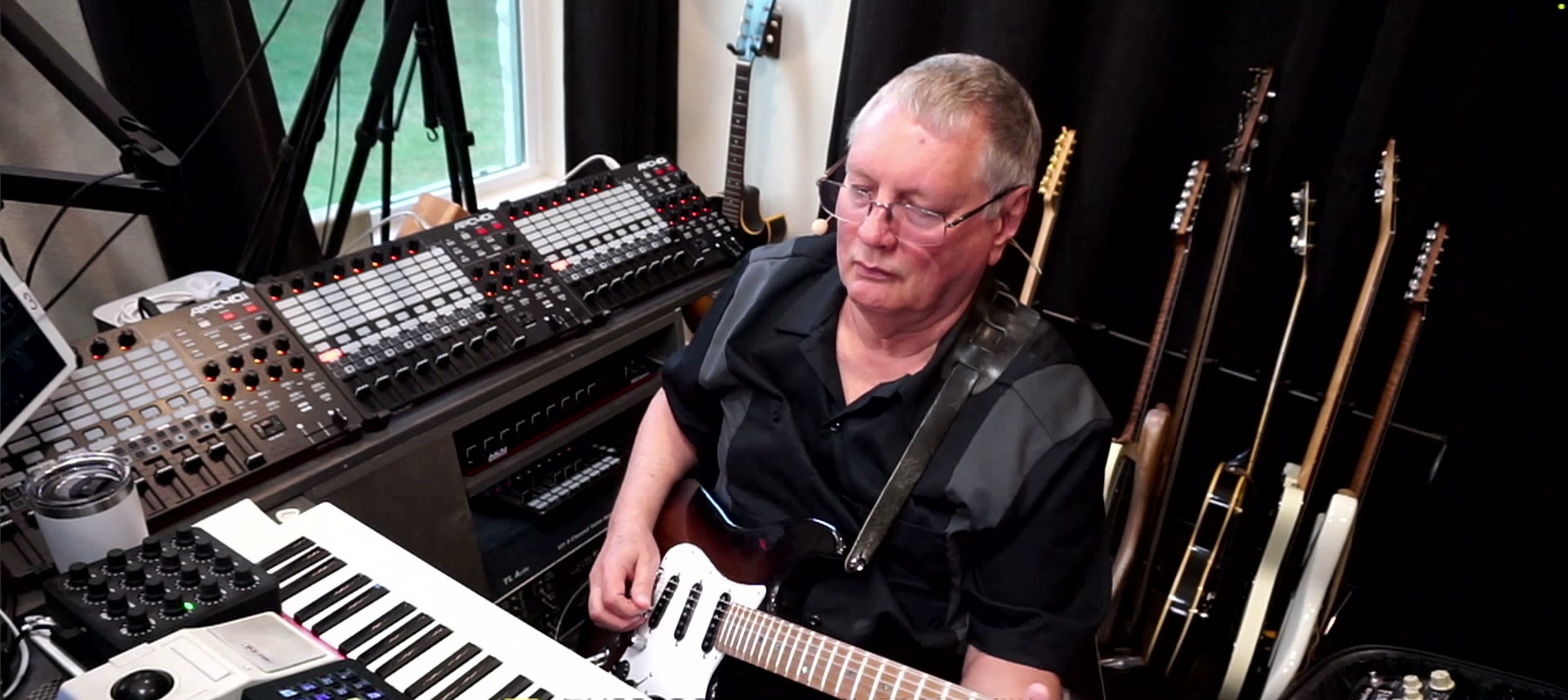 Larry Seyer playing guitar in his studio, surrounded by keyboards, controllers, and a wall of guitars